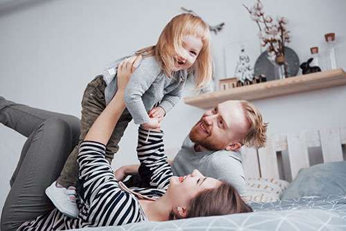 A mom and dad relaxing on their bed. Mom is holding their toddler daughter above her head, smiling.
