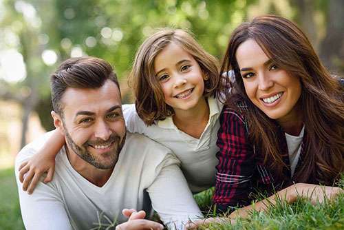 Mom, dad, and pre-teen daughter in a park, laying down on the grass, smiling at the camera.