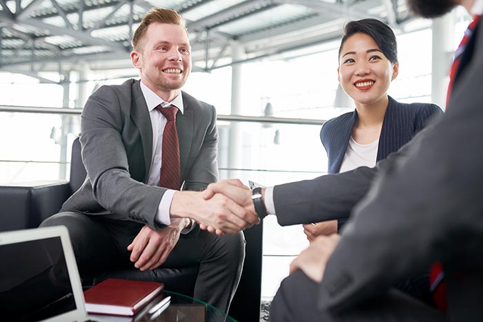 Three businesspeople sitting in an office creating a business partner buyout with whole life insurance  – a man is shaking another man’s hand as a smiling woman look on
