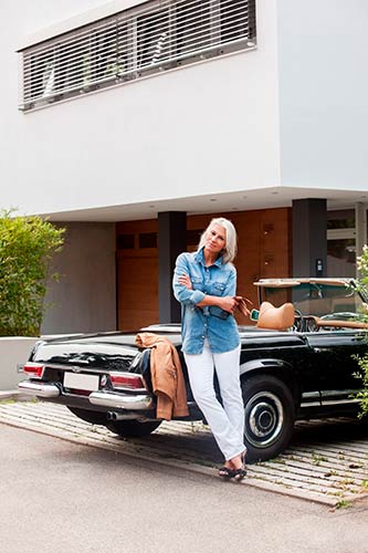 Happy middle-aged woman standing near her vintage car, in front of a swanky modern house.
