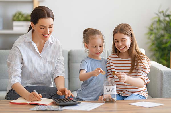 Mom working on a family budget while her two daughters add coins to a jar marked ‘save’