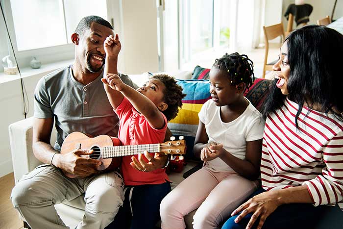 Family of a mom, dad, and two kids sitting on a couch as the dad plays a small guitar, all happy because they are protected by whole life insurance.