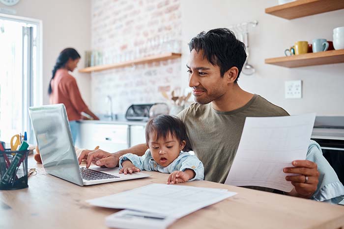 Man holds his baby while working on his laptop and looking at financial documents